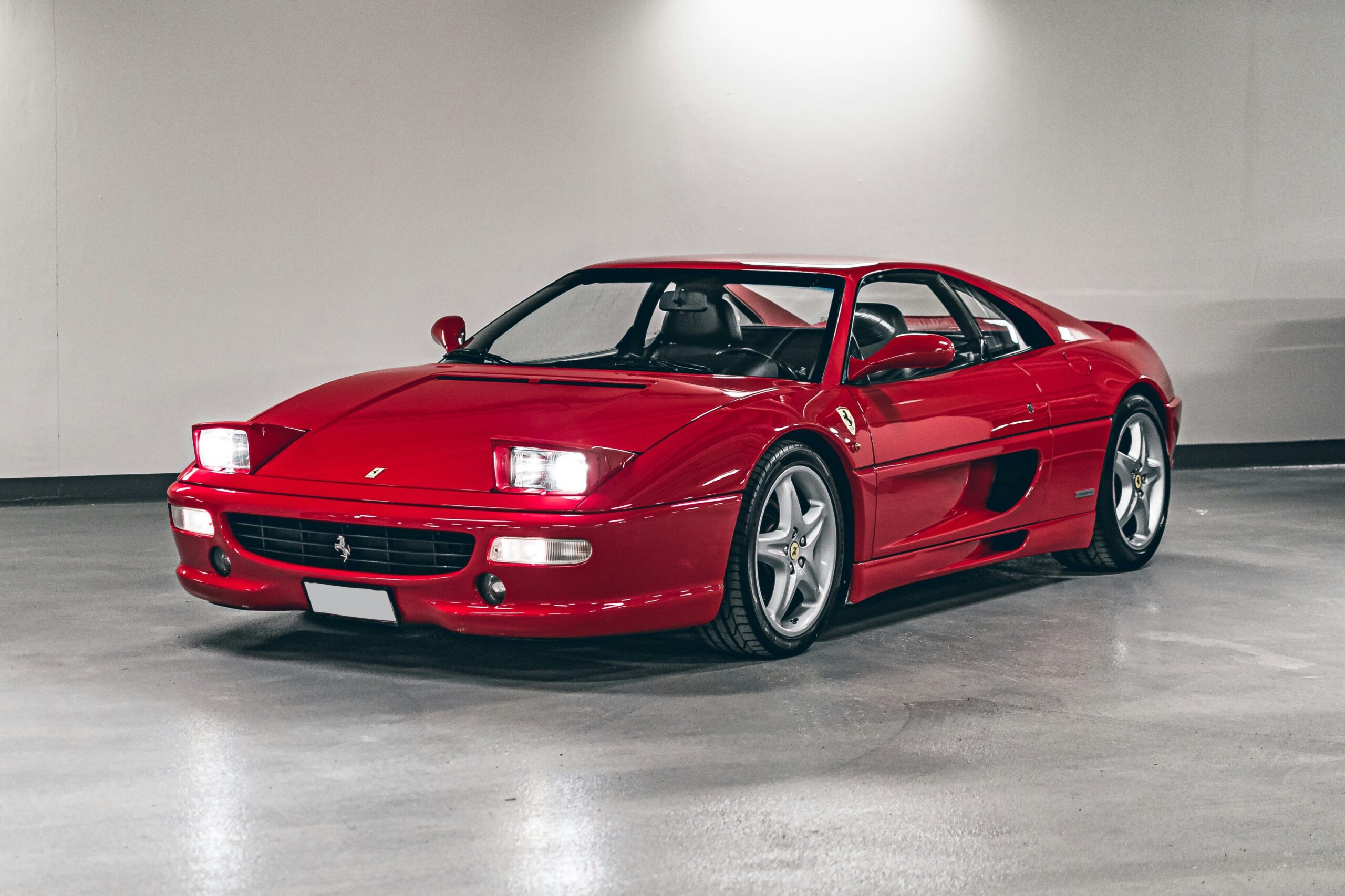 A red Ferrari F355 investment coupe parked indoors wth its headlights on.