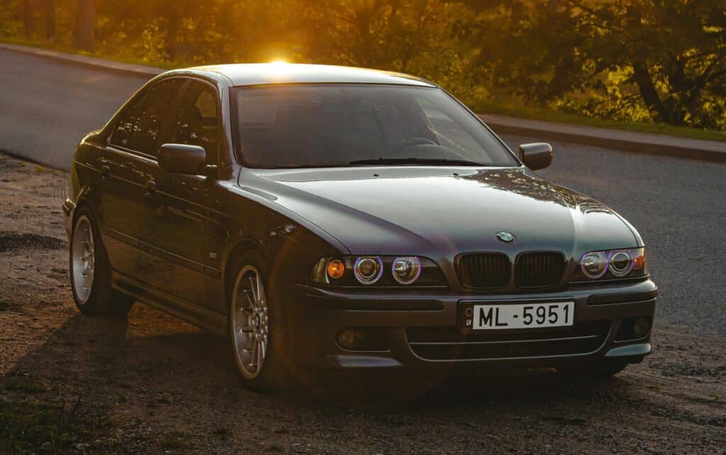 A silver BMW E39 M5 parked on a road with sun in the background.
